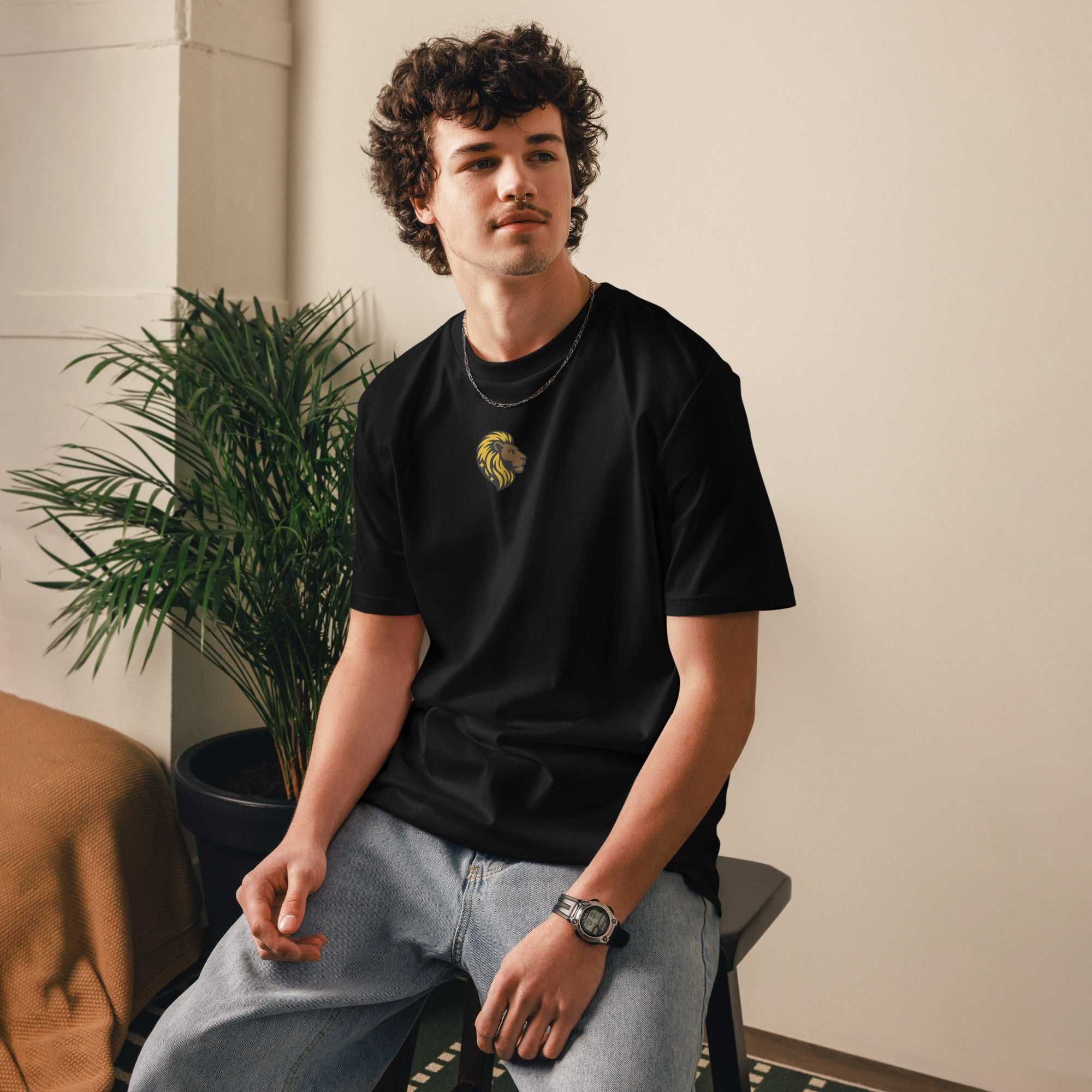 Young man wearing black He Is Risen T-Shirt with gold lion and cross design sitting indoors